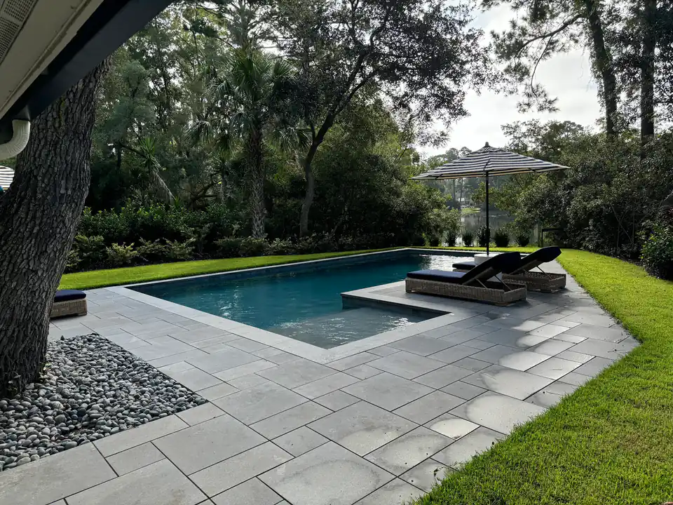 Rectangular pool under a live oak with gray pavers, a sun shelf and a striped umbrella