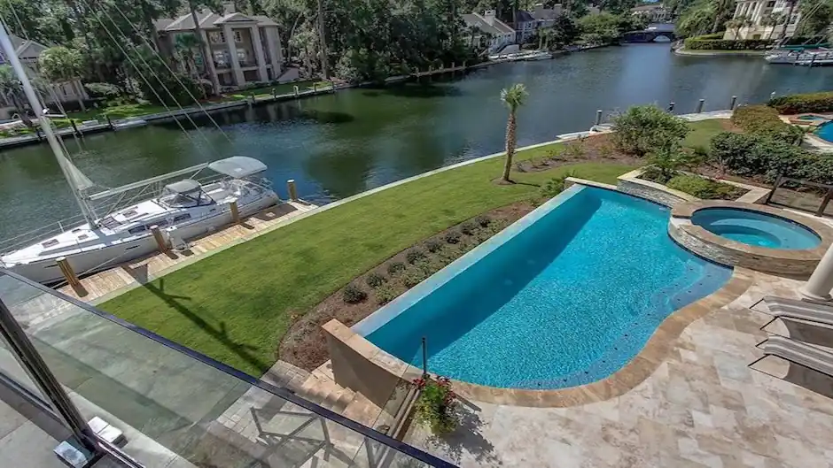 Aerial view of an infinity-edge pool and raised spa on a lawn beside a canal with a sailboat at the dock